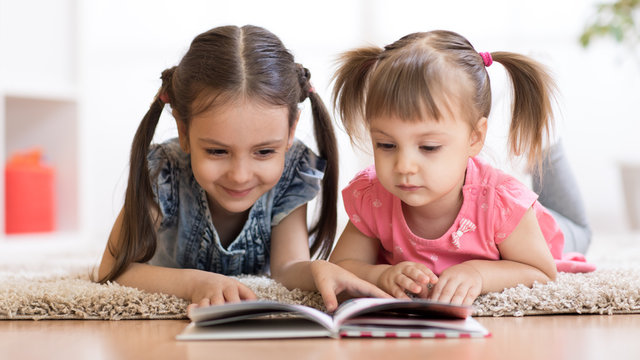 Child girl reading book with little sister at home