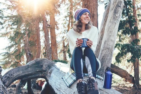 Traveling young woman with knitted hat drinking hot coffee from a mug and sitting on tree in wild forest at sunny day.