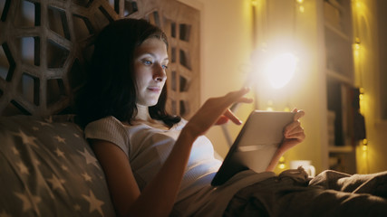 Close-up of young concentrated woman using tablet and watching online news lying in bed at home at night