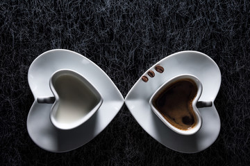 Two heart shaped cups with black coffee and milk pointing to each other on a black and silver kitchen table top in hard, low key light - 183452361