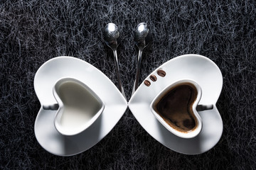 Two heart shaped cups with black coffee and milk pointing to each other in the form of a butterfly on a black and silver kitchen table top in hard, low key light - 183452392