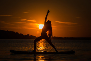 Silhouette of beautiful young girl exercising yoga on SUP in the scenic yellow sunset on lake Velke Darko, Zdar nad Sazovou, Czech republic