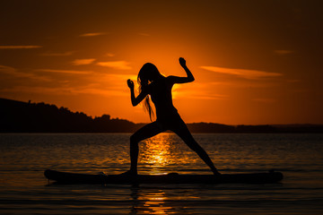 Silhouette of beautiful young girl exercising yoga on SUP in the scenic yellow sunset on lake Velke Darko, Zdar nad Sazovou, Czech republic