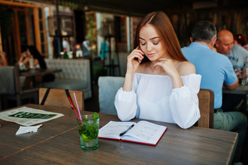 Portrait of a gorgeous young businesswoman talking on the phone and writing something down in her red notebook while sitting in a cafe.