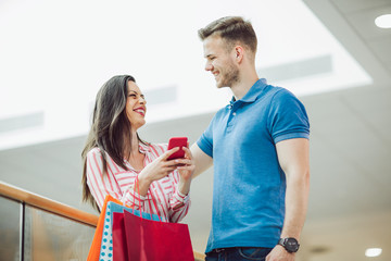 Fototapeta premium Happy young couple with shopping bags and smartphone talking in mall