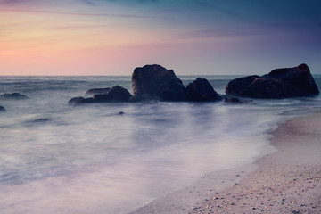 Summer seasonal natural vacation background. Romantic morning at sea. Big boulders sticking out from smooth wavy sea. Pink horizon with first hot sun rays. Long exposure.