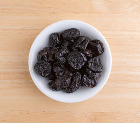Top view of a portion of dried bing cherries in a small white bowl atop a wood table.