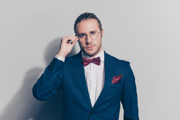 Portrait of stylish man holding with one hand glasses on face with serious expression in blue jacket, white shirt with red bow-tie and  pocket square, standing over grey background