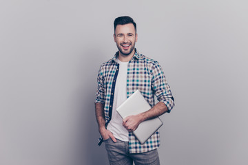 Portrait of smiling  guy in checkered shirt holding one hand in pocket and in another one  laptop, looking at camera, standing over grey background