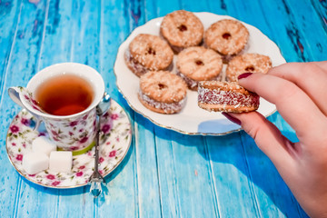 a plate with a shortbread cookie and a cup of black tea with pieces of sugar on a blue wooden background. A woman's hand holds a cookie. Vegetarian