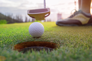 Golf ball putting by golf player in background, golf ball spining to the hold on the green of golf course with early light of sunset