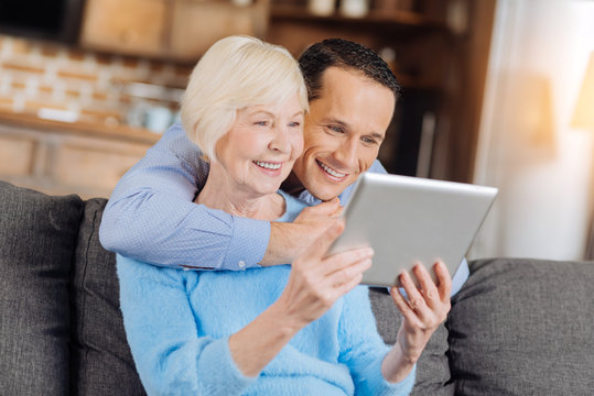 Amusing content. Charming young man hugging his mother from behind and watching a video on tablet together with her while both of them smiling brightly