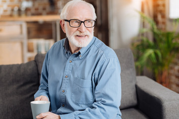 Favorite drink. Charming elderly man in eyeglasses sitting on the couch, holding a coffee cup and...