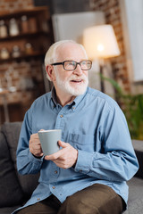 Healthy drink. Pleasant senior man in eyeglasses sitting on the sofa and drinking tea from the big mug while looking into the distance and smiling