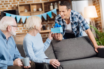 Pleasant surprise. Charming young man giving a birthday present to his elderly mother sitting on the sofa next to her husband