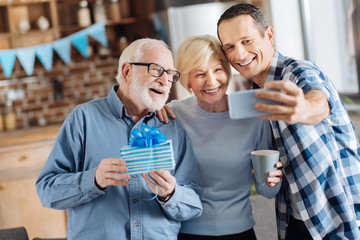 Seizing the moment. Cheerful young man taking selfies with his elderly parents during the celebration of the fathers birthday while the man holding a gift box