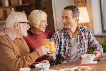 Nice morning. Pleasant senior woman hugging her son and husband while they having breakfast and drinking juice, clinking the glasses