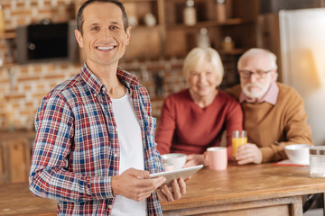 Favorite device. Charming young man standing near the kitchen counter and posing with a tablet while his elderly parents having breakfast in the background