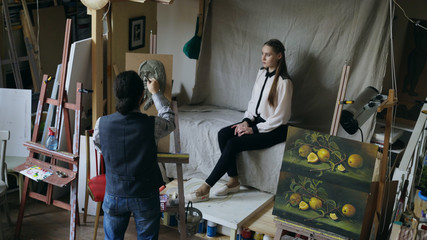 Fototapeta na wymiar Sculptor creating sculpture of human's face on canvas while young woman posing to him in art studio