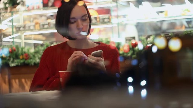 Beautiful young girl using smartphone sitting in cafe behind window. Xmas time