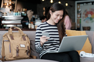 Beautiful asia girl using laptop computer and smart phone while sitting on sofa with smiling face emotion, people and technology concept, lifestyle