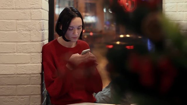 Pretty young girl using smartphone in cafe in evening. Christmas time