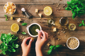 Woman hands making italian pesto in bowl. Ingredients - basil, lemon, parmesan, pine nuts, garlic, olive oil and salt on rustic wooden background. Top view, flat lay, copyspace