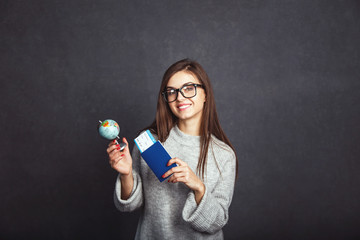 Cheerful girl holding passport, plane ticket and globe before grey background, indoor travel concept