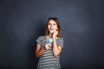 Cheerful girl holding passport, plane ticket and globe before grey background, indoor travel concept