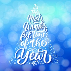 Custom lettering It's the most wonderful time of the year in Christmas fir tree silhouette illustration, on winter blue defocused background. Vector holiday poster design. © Ann Lou