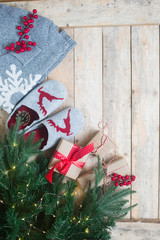 Christmas gifts concept, home grey slippers with red raindeer, warm sweater snowflake pattern, gift boxes red ribbon twine pine branches hot coffee marshmallow candy canes berries wooden background