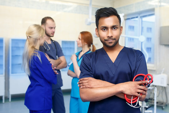 Asian Male Doctor In Front Of Team, Looking At Camera With Medical Team In Background. Multiracial Team Of Young Doctors In A Hospital Standing In A Operating Room.