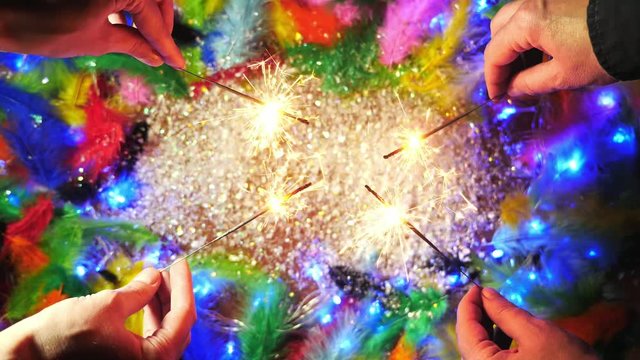 The hands lighting sparklers on the background with colorful feathers, white snow and blinking garland.Top view