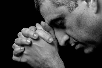 Close up of faithful mature man praying, hands folded in worship to god with head down and eyes closed in religious fervor. Black background. Concept for religion, faith, prayer and spirituality. - obrazy, fototapety, plakaty