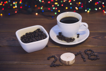 A romantic festive still life with a cup of coffee with a saucer, a bowl of coffee beans and 2018 inscription of coffee beans on a rustic wooden background. dark colorful bokeh