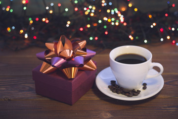 A romantic festive still life with a cup of coffee with, coffee beans on a saucer and a burgundy gift box with an orange-purple bow on a rustic wooden background. Dark colorful bokeh