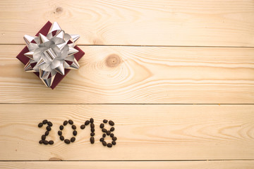 A light wooden Christmas or New Year background with 2018 inscription of coffee beans and a burgundy gift box with a silver bow. Top view. Copy space