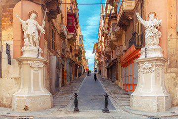 The traditional Maltese street stairs with corners of houses, decorated with statues of saints St. John and St. Paul and building with colorful balconies in Valletta, Capital city of Malta - obrazy, fototapety, plakaty