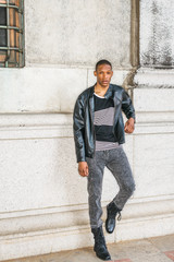 Man Urban Casual Fashion. Wearing black leather jacket, striped undershirt, jeans, leather boots, a young African American guy standing against wall on street in New York, relaxing, waiting for you..