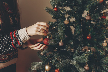 woman in stylish sweater decorating christmas tree in room. merry christmas and happy new year concept. space for text. woman putting on red ornaments. family cozy moments
