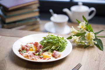 Breakfast at the table by the window. Fried eggs with bacon and herbs, hot tea in a teapot and a cup. Books, fresh flowers, bright sun from the window.