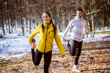 Pretty smiling flexible girl in sportswear doing leg stretching with her personal trainer in the forest in the sunny winter morning.