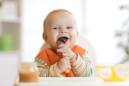 Cheerful baby child eats food itself with spoon. Portrait of happy kid boy in high-chair.