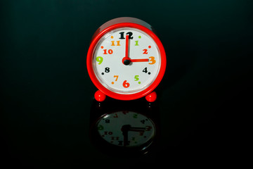 Red alarm clock with time shows 3 o'clock with reflections on an isolated black background.
