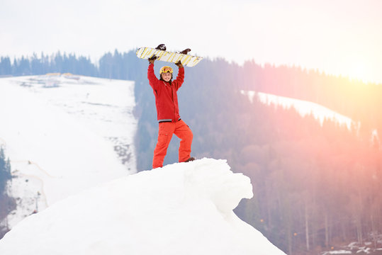 Young man snowboarder standing on the top of the snowy slope with snowboard in hands above a head, smiling to the camera in the evening at sunset. Skiing and snowboarding concept