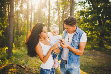 stylish young Family mom, dad and daughter one year old blonde sitting with father on shoulders playing happy and smiling, outdoors outside the city in the woods in summer at sunset.