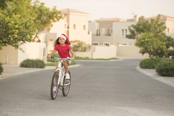 A biracial child wearing Santa hats while riding her bike on a street in a neighbourhood with homes in the background - Powered by Adobe
