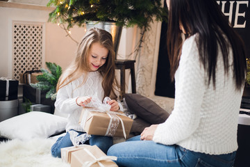 Merry Christmas and Happy Holidays. Cheerful mom and her cute daughter girl opening a Christmas present. Parent and little child having fun near Christmas tree indoors