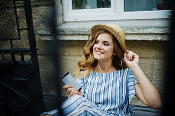 Portrait of a gorgeous model in striped overall sitting on the chair outdoor and listening to music in earphones.