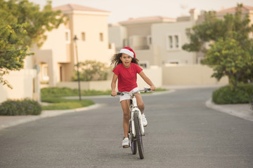 A biracial child wearing Santa hats while riding her bike on a street in a neighbourhood with homes in the background